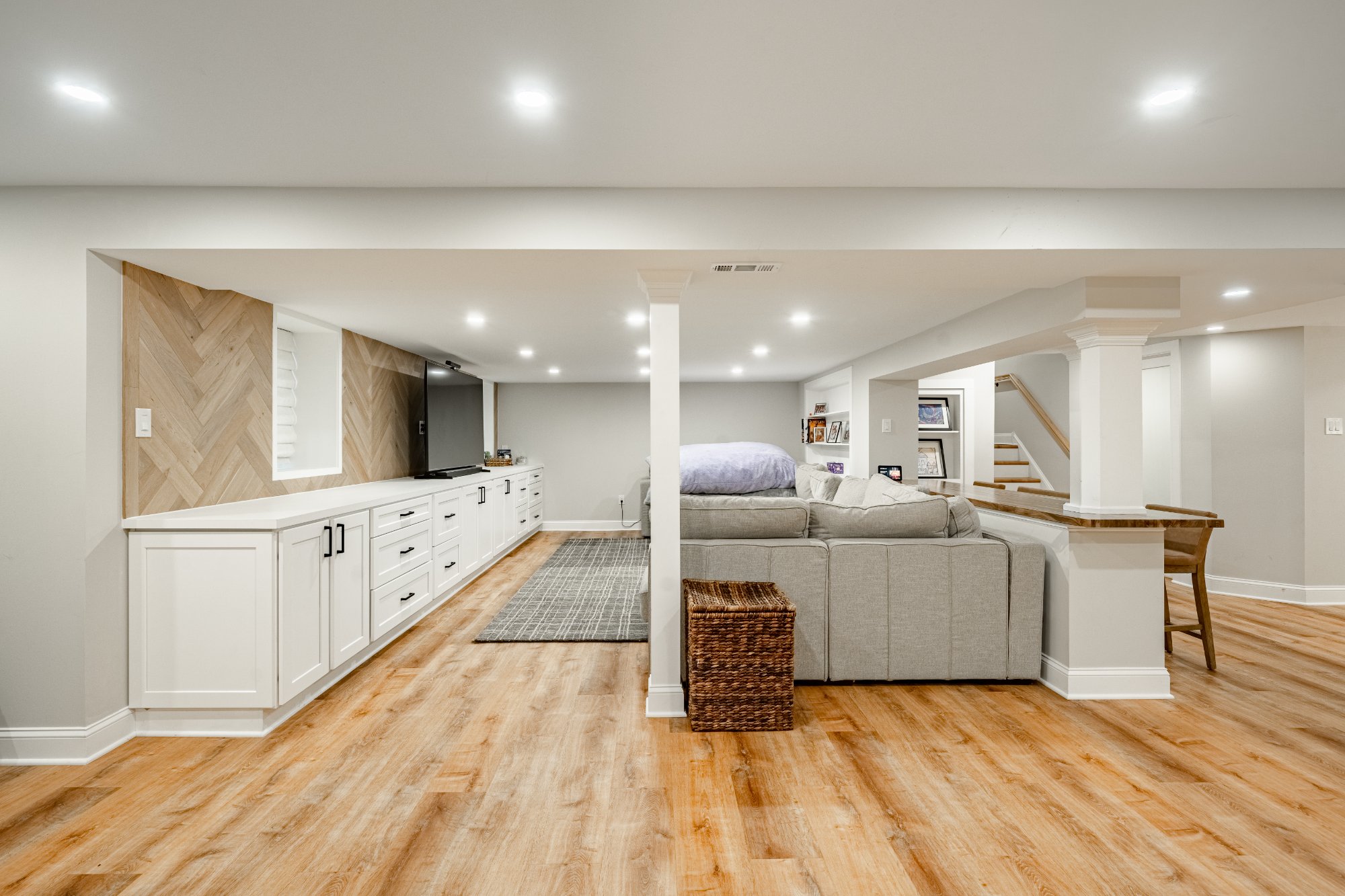 Finished basement living space with herringbone accent wall, white built-in cabinetry, LVP flooring, and open staircase — Saratoga Drive project