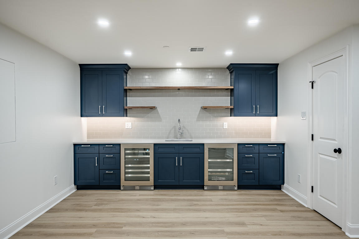 Plymouth Road wet bar — navy shaker cabinetry, white subway tile backsplash, wood floating shelves, dual wine refrigerators, quartz countertops