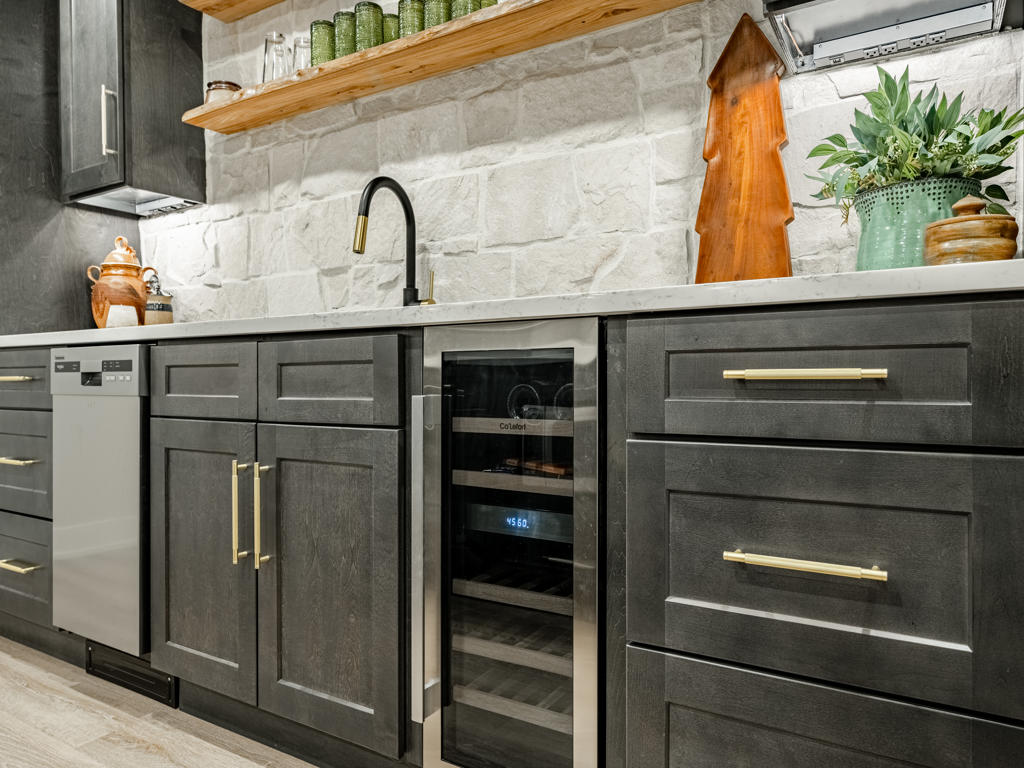 Close-up of basement wet bar lower cabinetry with built-in wine cooler, stainless dishwasher, and brushed brass hardware on charcoal shaker doors