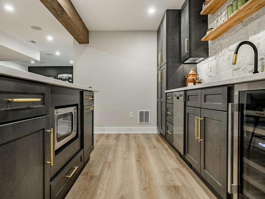 Corridor view between kitchen island and wet bar wall showing brass hardware detail, microwave drawer, wine cooler, and stone backsplash
