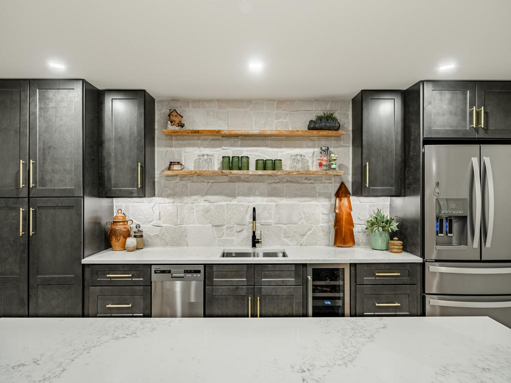 Full basement bar wall with charcoal shaker cabinets, floating wood shelves on natural stone, undermount sink, wine cooler, and stainless French door refrigerator