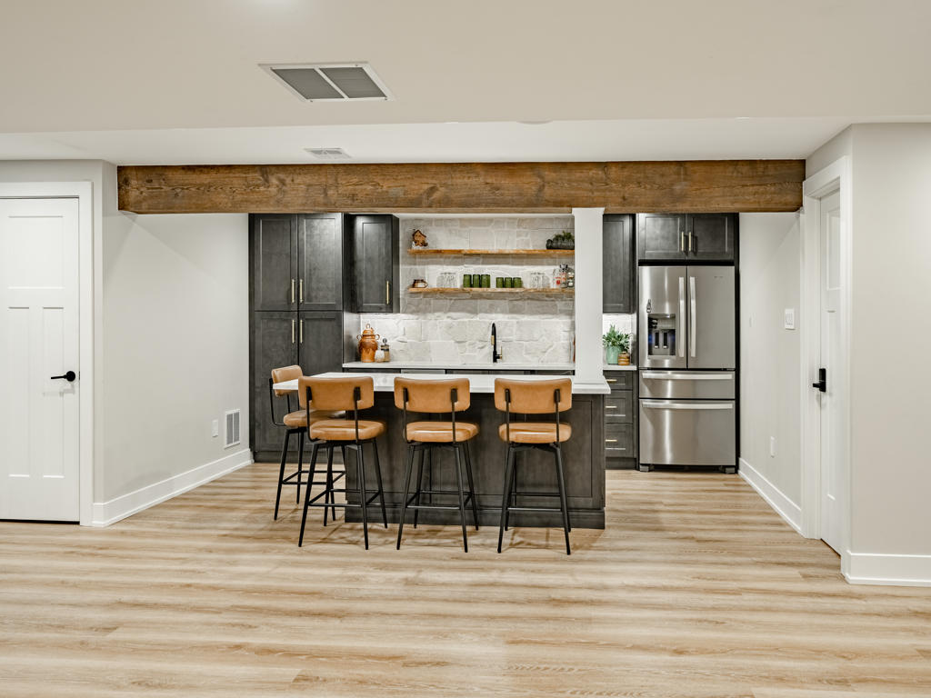 Basement wet bar and kitchenette from entry side showing reclaimed beam overhead, cabinetry, stone backsplash, and saddle leather bar stools