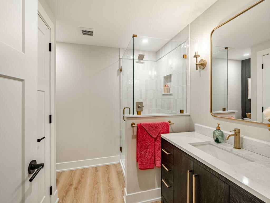 Full basement bathroom in Chester Springs PA with dark wood vanity, white quartz countertop, gold-framed mirror, brass sconces, and frameless glass shower