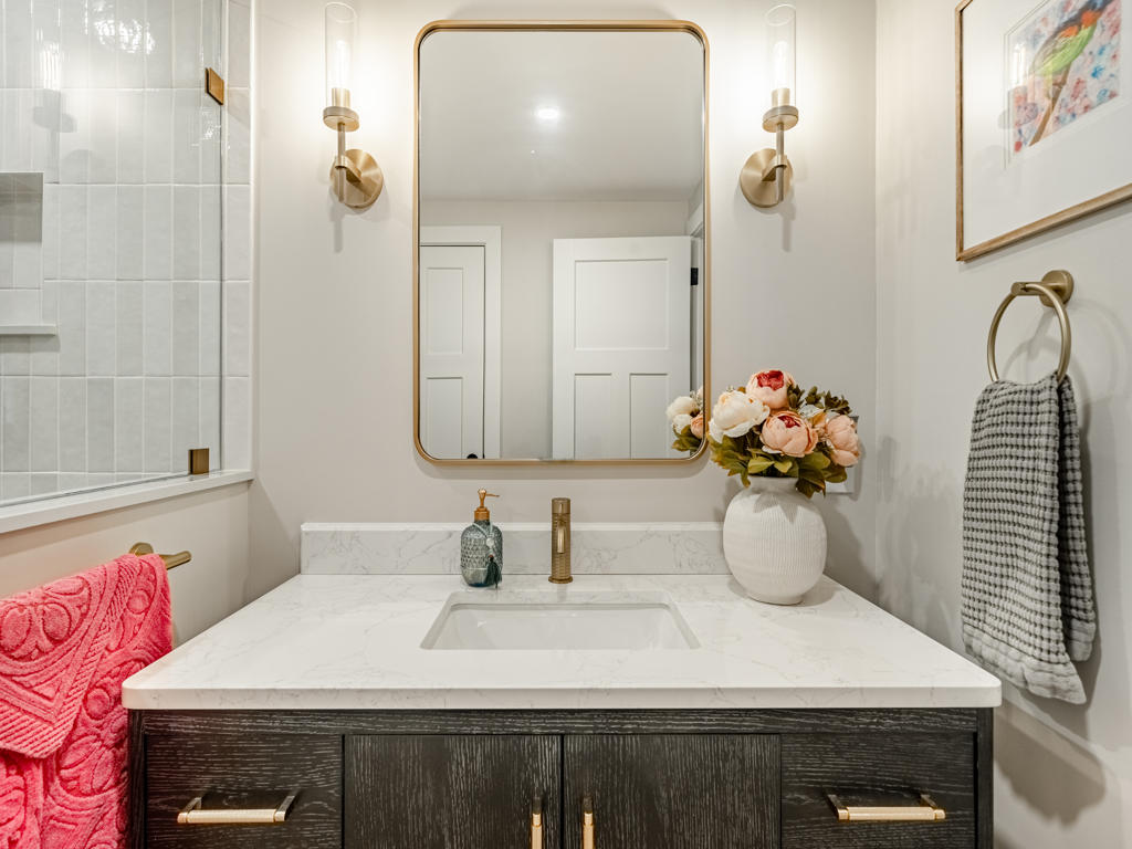 Basement bathroom vanity close-up with dark espresso wood, quartz countertop, brass faucet, gold-framed mirror, and brass cylinder wall sconces