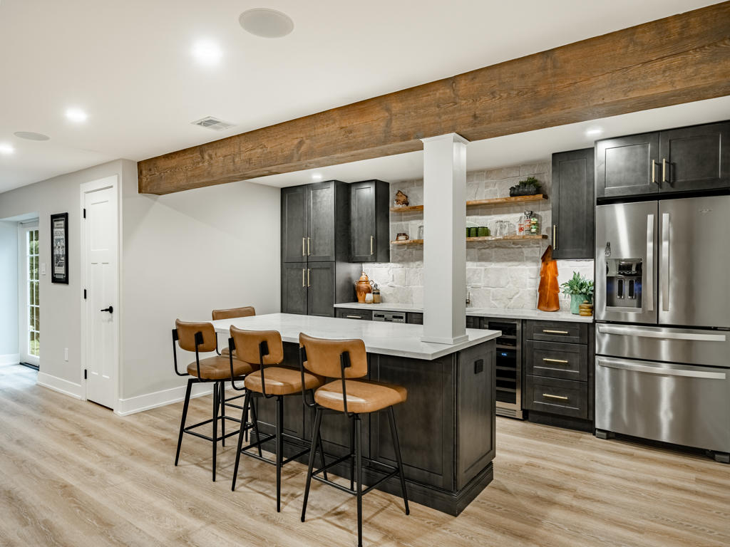 Custom basement wet bar in Chester Springs PA featuring charcoal shaker cabinets with brass hardware, black walnut island, natural stone backsplash, and reclaimed wood ceiling beam