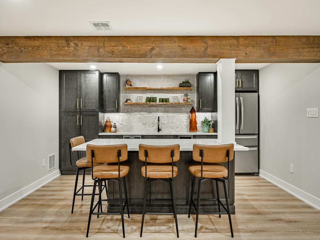Straight-on view of basement wet bar with charcoal shaker cabinets, natural ledger stone backsplash, floating wood shelves, and white quartz countertops