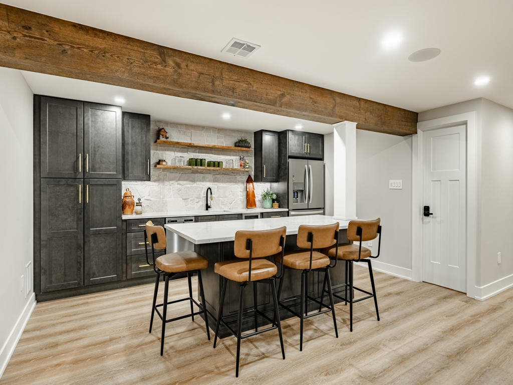 Basement kitchenette with charcoal cabinetry, natural stone backsplash, black walnut bar top, stainless refrigerator, and reclaimed barn wood beam overhead
