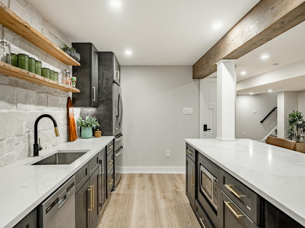 Galley view of basement wet bar showing stone backsplash, floating wood shelves, island microwave drawer, brass hardware, and reclaimed ceiling beam