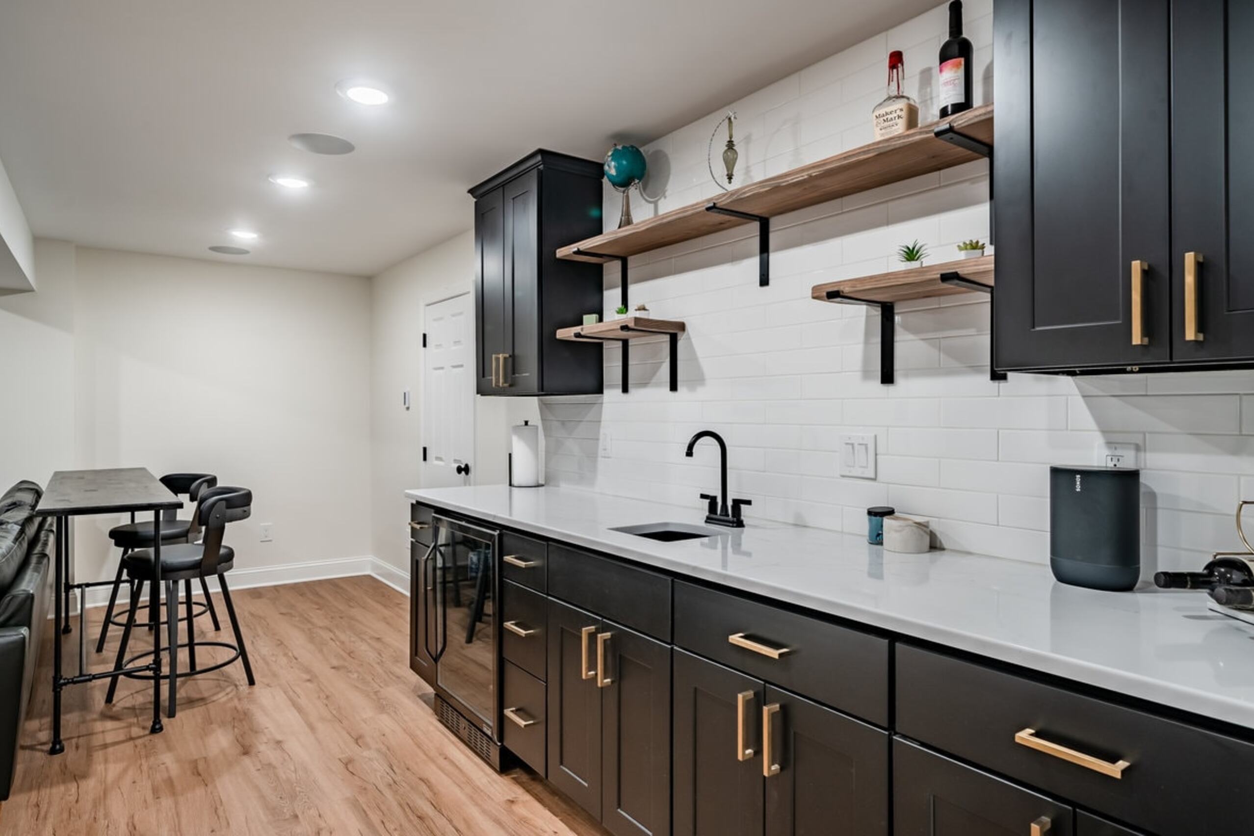 Chester Springs basement wet bar with black shaker cabinets, gold hardware, floating wood shelves, and white subway tile