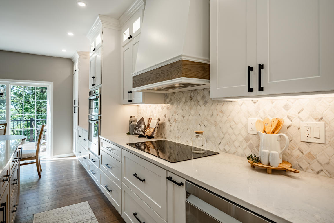 Cooking wall showing custom wood-banded hood over induction cooktop, arabesque backsplash, white shaker cabinets with matte black hardware, and stacked ovens beyond