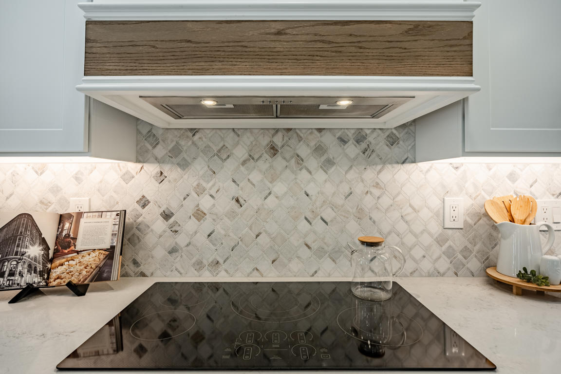Close-up of induction cooktop with glass surface flush-mounted in quartz countertop, wood-banded range hood above, arabesque marble backsplash