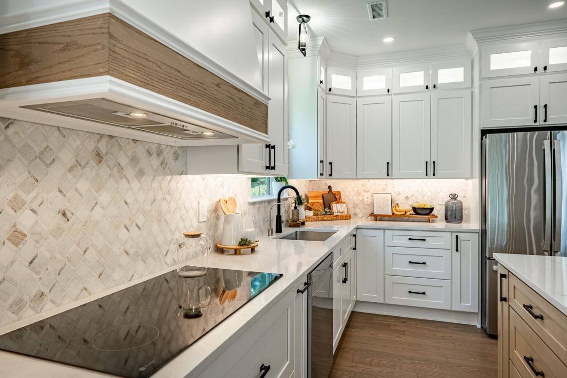 Kitchen corner view showing wood-banded hood, induction cooktop, arabesque backsplash, sink area, natural oak island, and stainless refrigerator