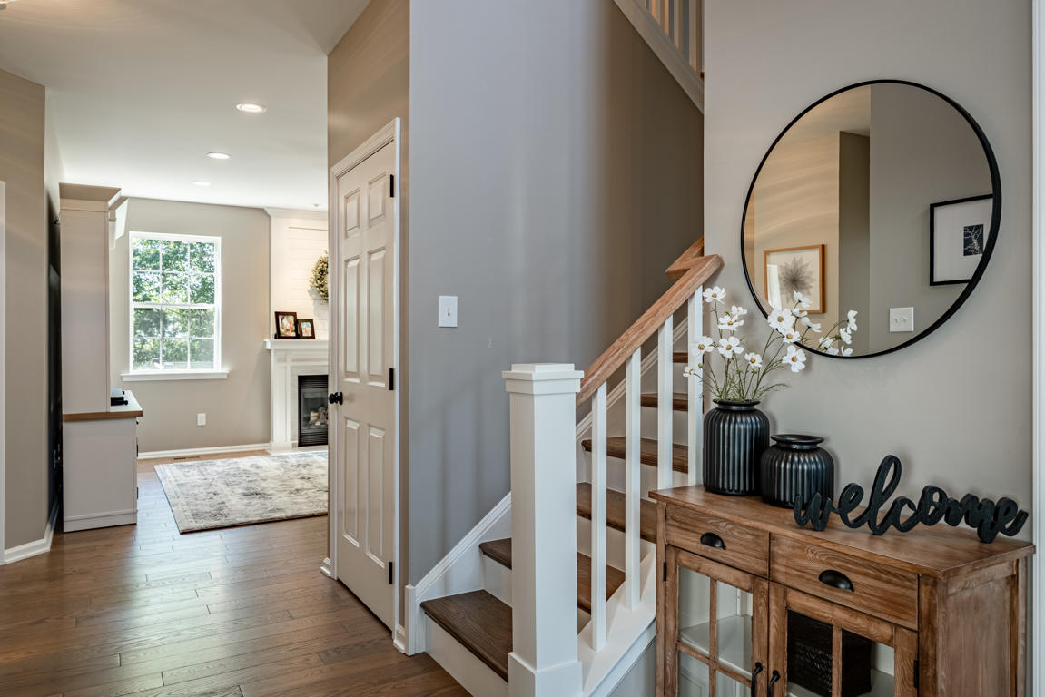 Entry console area with rustic oak cabinet, black decorative vases with white flowers, welcome sign, round black-framed mirror, and staircase beyond