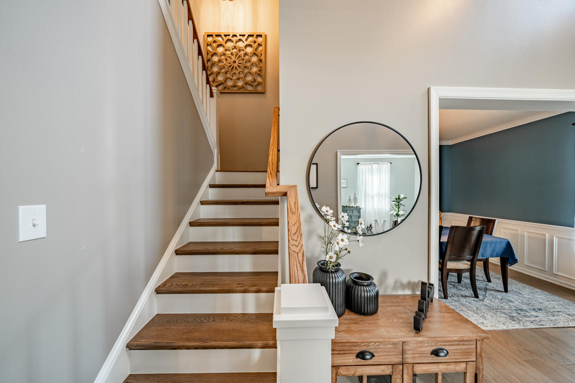 Staircase landing with carved wood wall art, stained oak treads, white risers, round mirror, entry console, and dining room doorway with blue accent wall