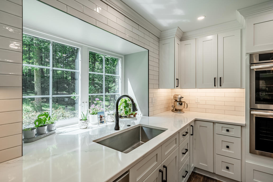 Corner sink area with large picture window, subway tile to ceiling, and white shaker cabinetry