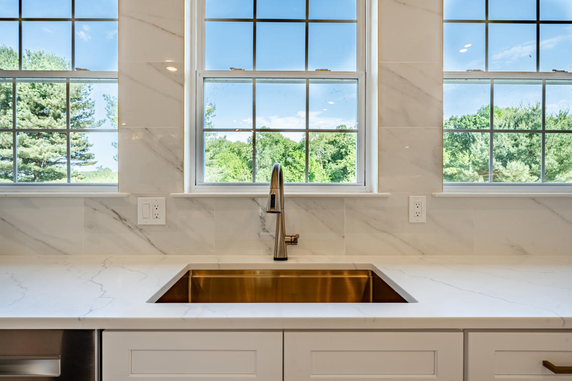 Gold undermount sink with pull-down sprayer faucet centered below three windows with marble-look backsplash and quartz countertop