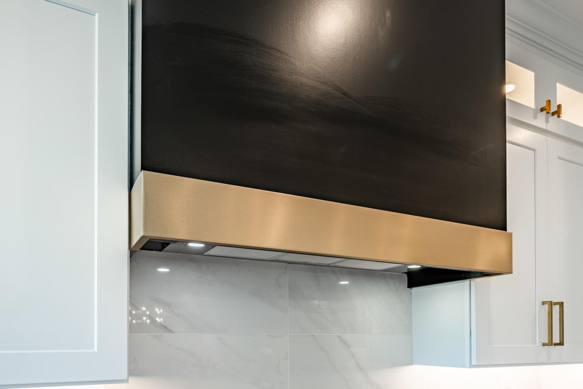 Matte black range hood close-up showing brushed gold accent band, recessed lighting, and marble tile backsplash