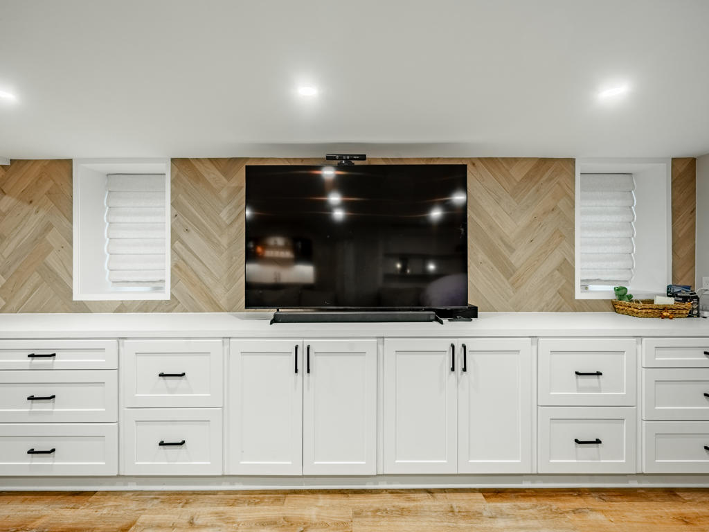 Close-up of white oak herringbone accent wall with large format TV, flanking egress windows, and white shaker media cabinetry with black hardware and quartz top