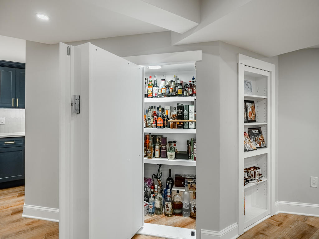 Hidden bookshelf door open in West Chester PA basement revealing fully stocked liquor storage closet with interior lighting, with second bookshelf door visible beside it
