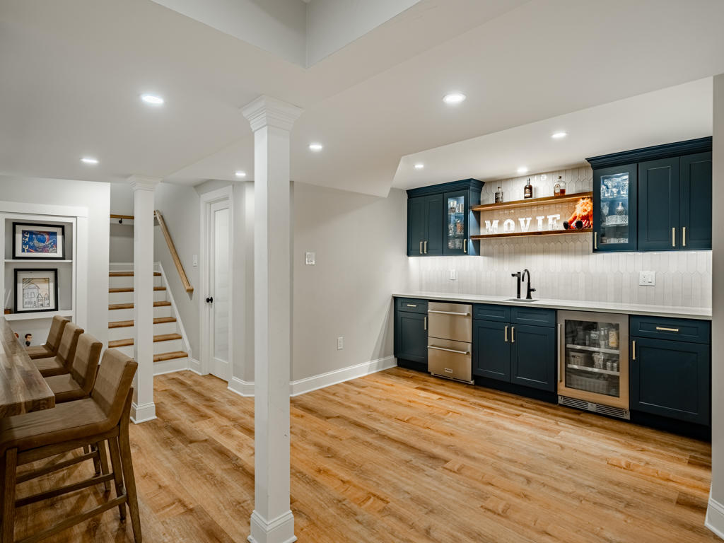 West Chester basement bar area viewed from seating zone showing navy cabinetry with gold hardware, bar stools, staircase, and built-in shelving