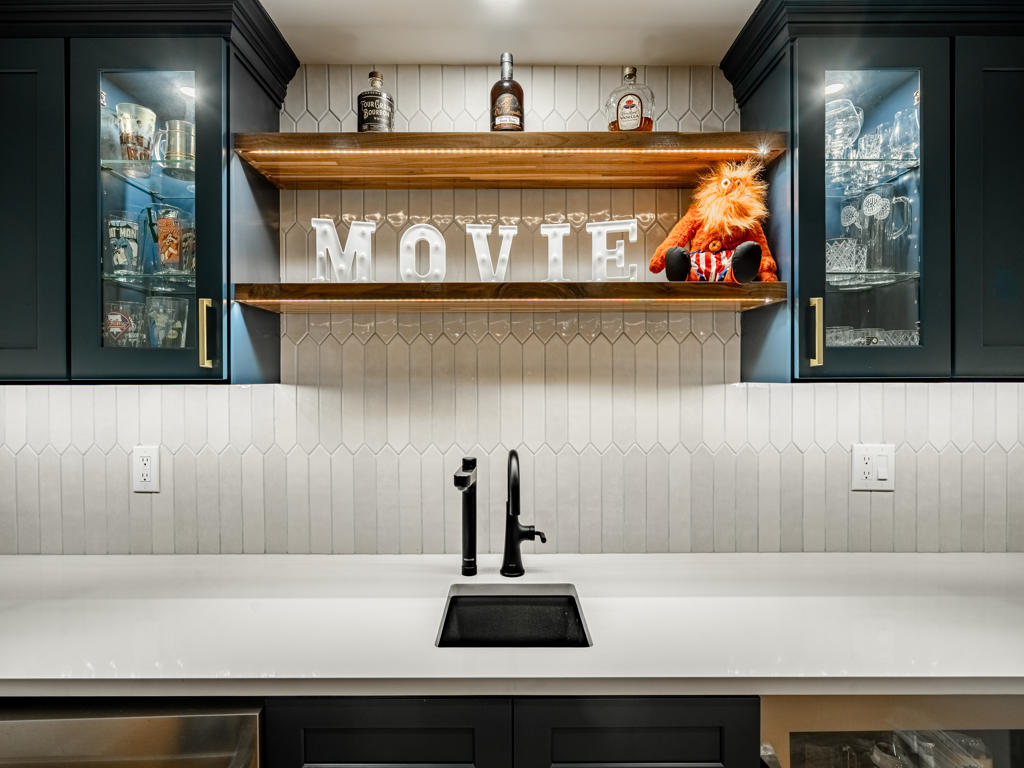 Close-up of navy bar zone with matte black undermount sink, black pull-down faucet, white quartz countertop, floating wood shelf, and lit glass-front cabinets in West Chester basement
