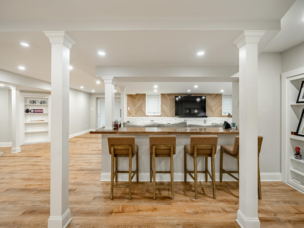 Wide angle view of finished West Chester PA basement remodel featuring custom white built-in media cabinetry, butcher block bar counter with leather stools, white oak herringbone TV accent wall, and open entertainment space with decorative columns