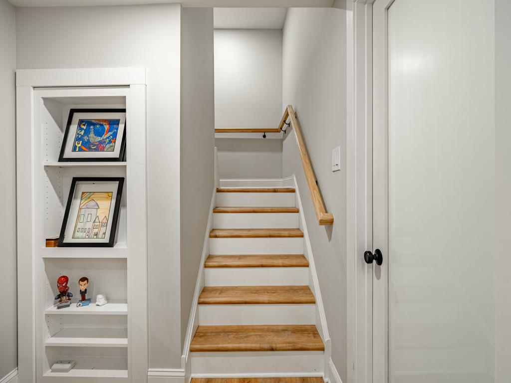 Finished basement staircase in West Chester PA with white risers, natural oak LVP treads, wood handrail, and custom built-in shelving niche beside stairs