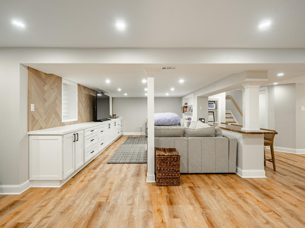 West Chester basement remodel wide view showing LVP flooring, white media cabinetry, herringbone accent wall, sectional seating, and open floor plan with decorative columns