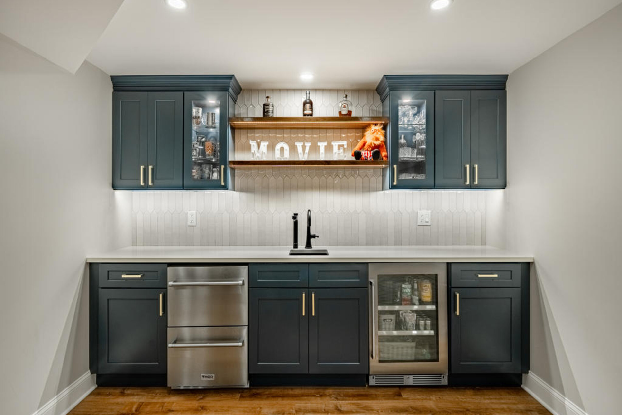 Navy shaker wet bar with Thor appliances, glass-front cabinets, and live-edge floating shelf in West Chester basement