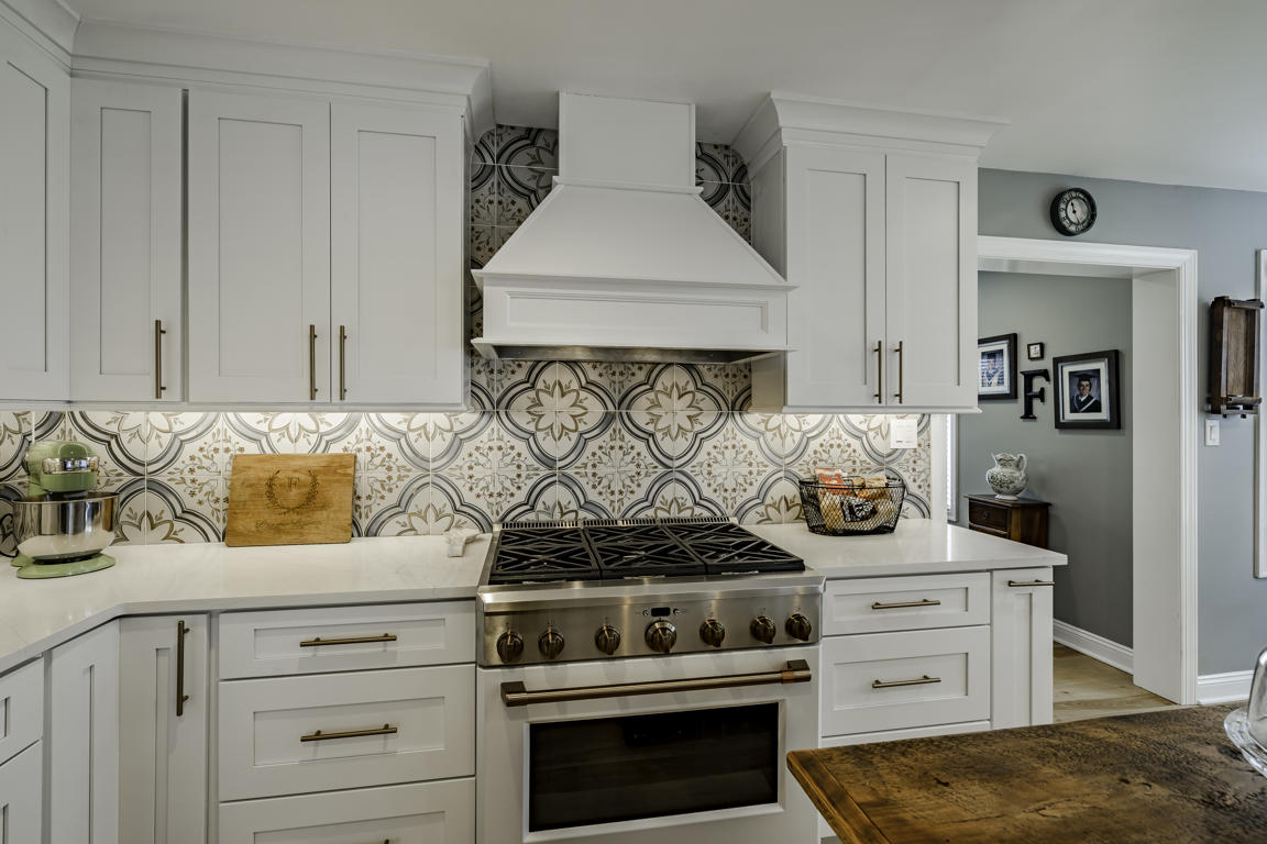 Custom white paneled range hood above stainless gas range with decorative cement tile backsplash and quartz countertops