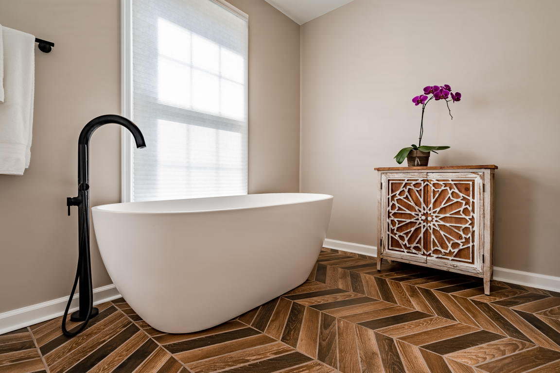 Freestanding soaking tub with floor-mounted black faucet beside Moroccan accent cabinet with orchid under tall window