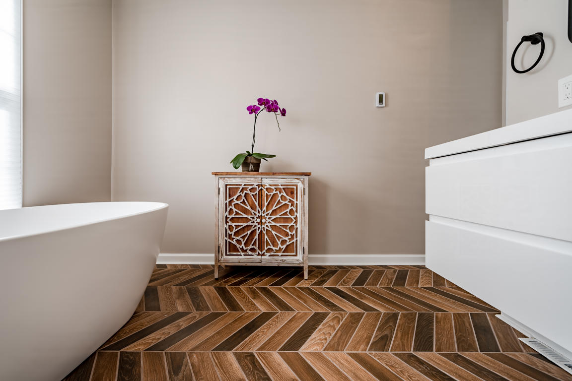 Tub area wide view showing freestanding soaking tub, accent cabinet, floating vanity, and chevron floor tile throughout