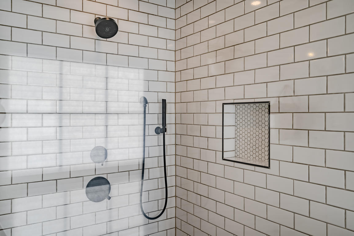 Shower interior detail showing white subway tile walls, matte black handheld shower wand, and penny round mosaic niche